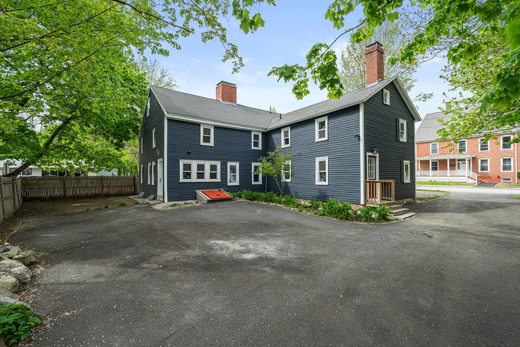 Wide view of the dark-clad two-story property with two chimneys, leafy trees, and a broad paved area.