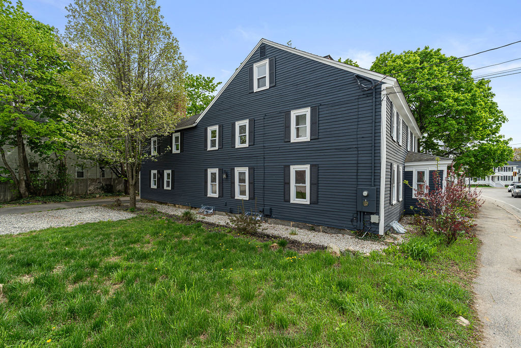 Oblique roadside view of the dark-clad property with many windows, gravel area, lawn, landscaping, and an intersection beyond.