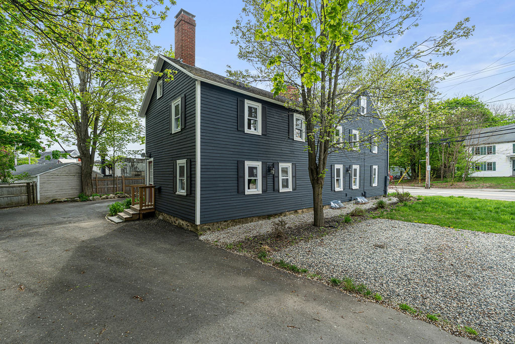 Corner exterior view of the dark-clad property with a chimney, mature trees, gravel area, and paved drive.