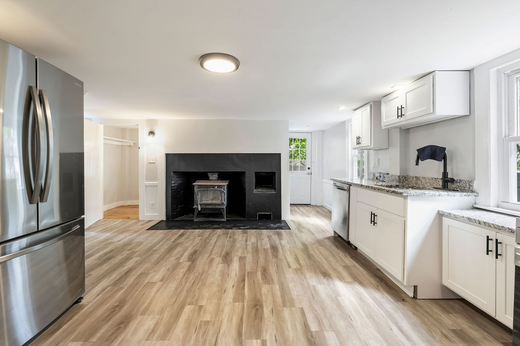 Long kitchen with stainless appliances, white cabinetry, stone-pattern counters, plank flooring, and a wood stove.